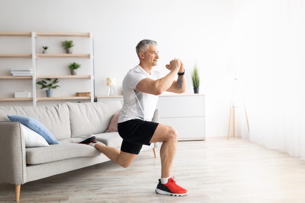 Active mature man doing squats while training at home, doing exercises near sofa in living room interior, keeping fit during covid-19 quarantine, empty space