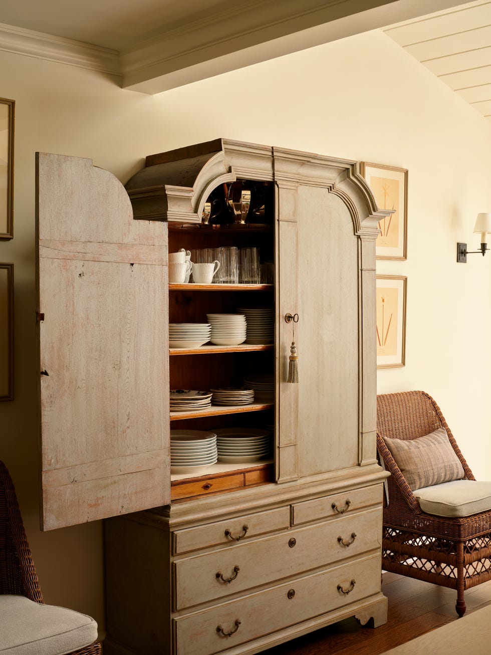 Antique wooden cabinet with open doors displaying plates and glasses, alongside a wicker chair in a cozy interior setting.