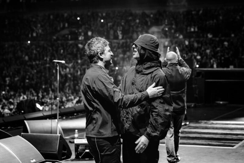 Two men stand on a concert stage facing each other, with one man wearing a bucket hat and the other placing a hand on his shoulder.