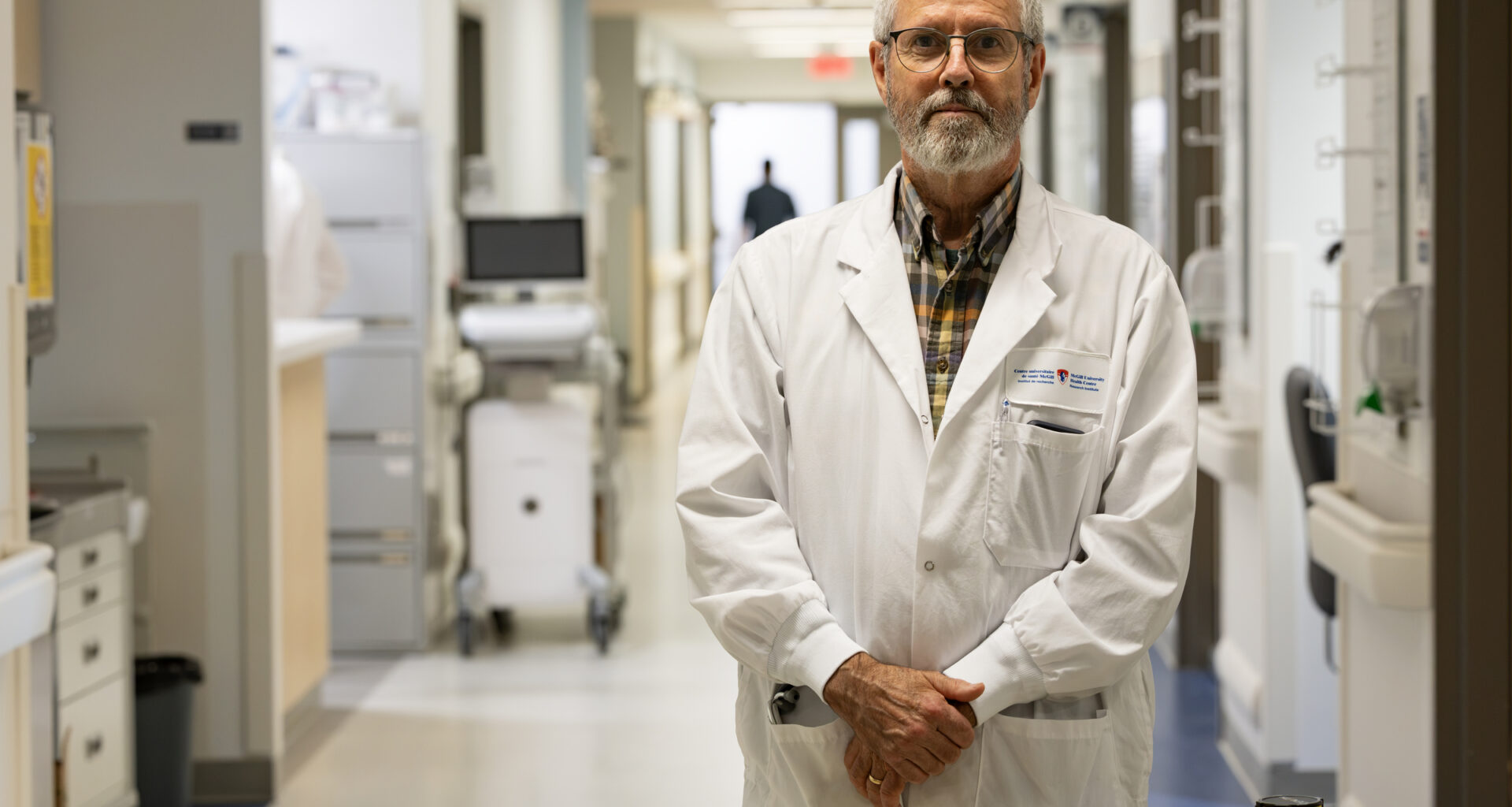 Dr. Brian Ward, in a white lab coat, poses for a photo standing in a medical centre hallway behind a barrier reading Attention ne pas entrer.