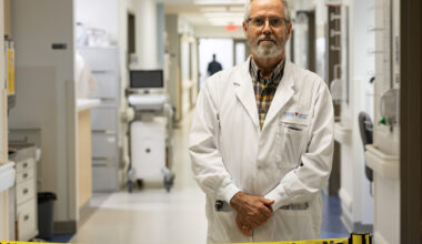 Dr. Brian Ward, in a white lab coat, poses for a photo standing in a medical centre hallway behind a barrier reading Attention ne pas entrer.
