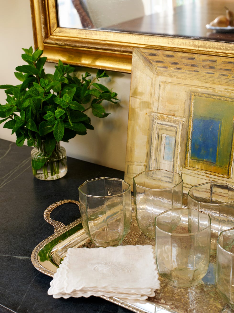 A silver tray with clear glasses, a folded napkin, a small plant, and a framed artwork reflected in a mirror.