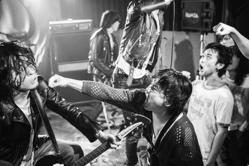 A black-and-white shot of a band performing on stage, with a guitarist shouting while another band member stands nearby.