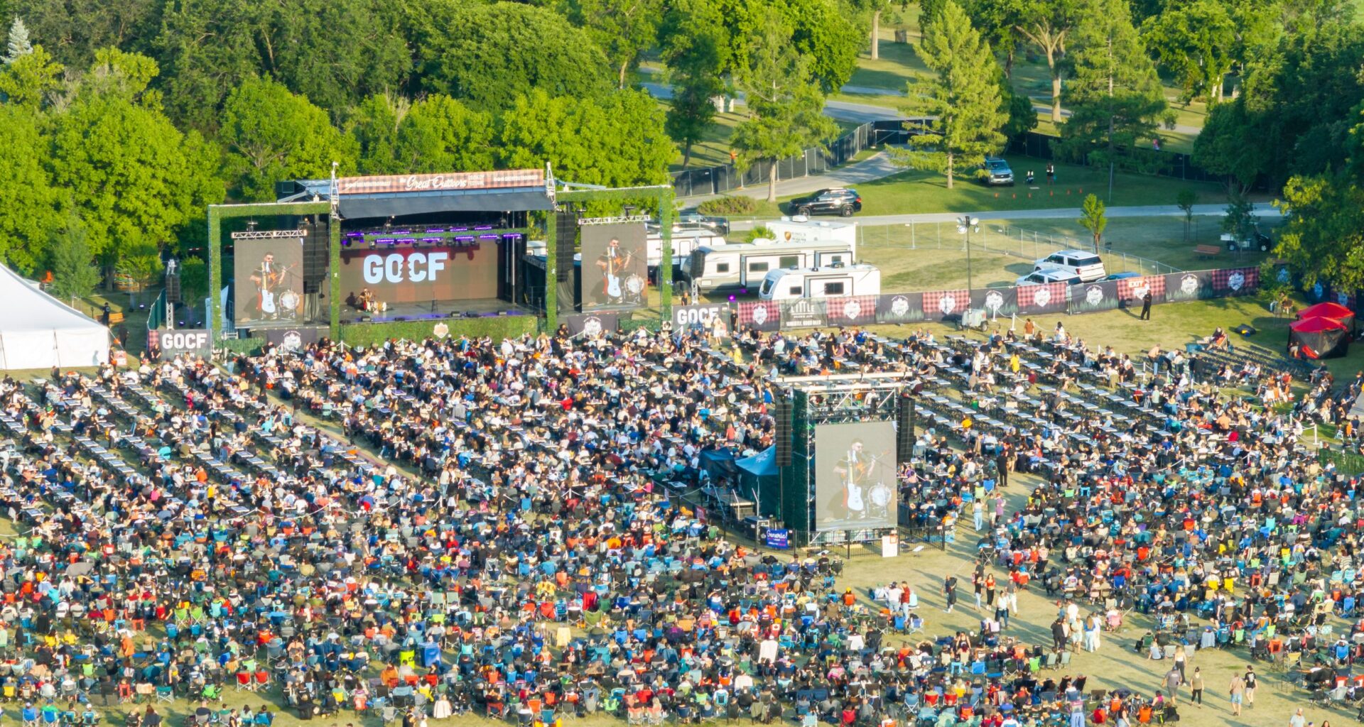 Crowd of people in an outdoor setting within trees and near a stage.