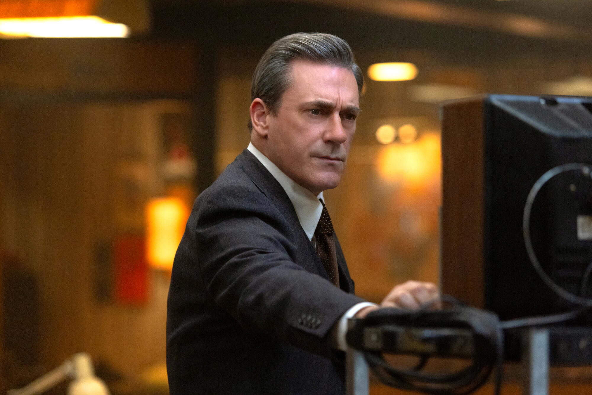 A man in a suit looks at a TV screen, leaning his hand against the cart it is on.