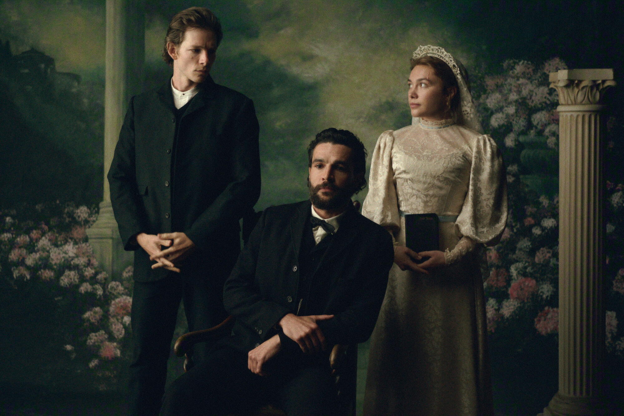 A man in a dark suit and a woman in a white bridal gown stand behind a man in a dark suit sitting in a chair.