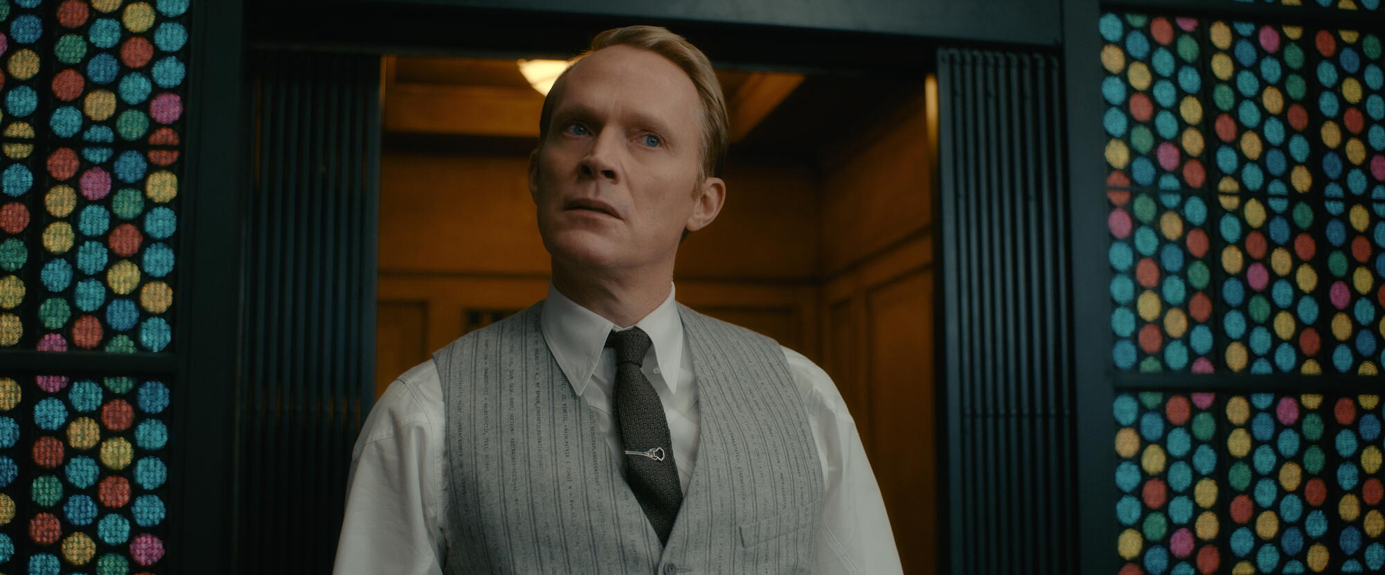 A man in a vest and tie standing in a doorway near walls covered in blue, green, yellow and red dots.
