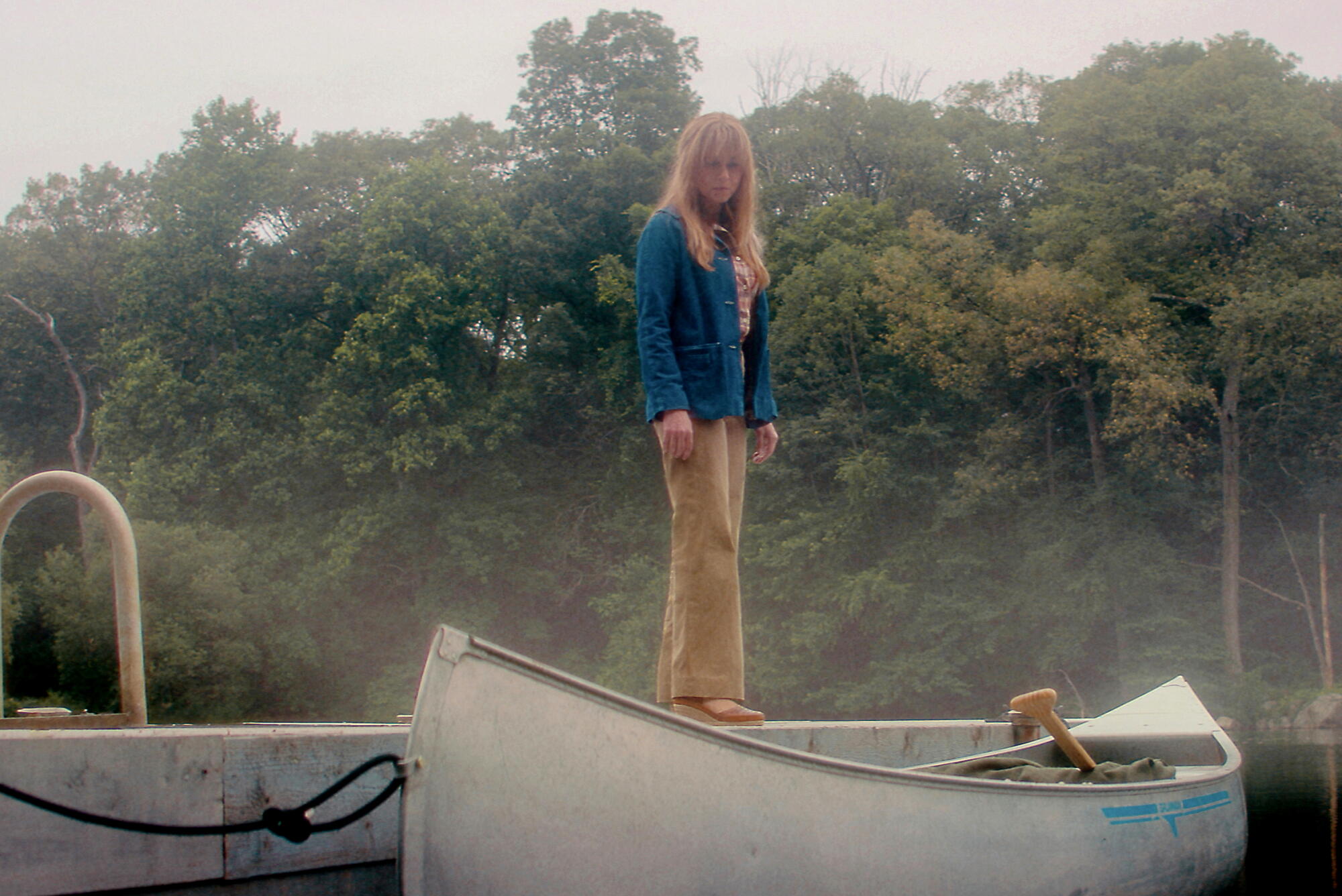 A woman in a blue shirt and khaki pants stands on a dock near a canoe on a foggy lake.