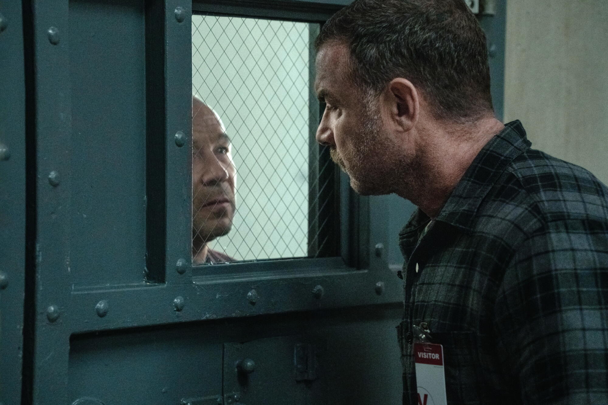 A man looks through the window of a steel door as another man looks back at him from the other side.