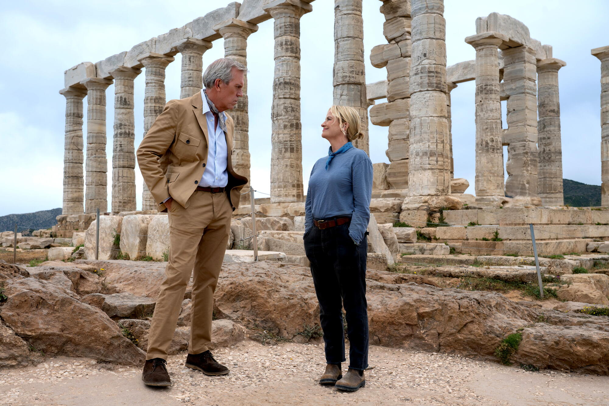 A man in a khaki jacket and pants stands next to a woman in blue shirt and pants on the site of ancient ruins.