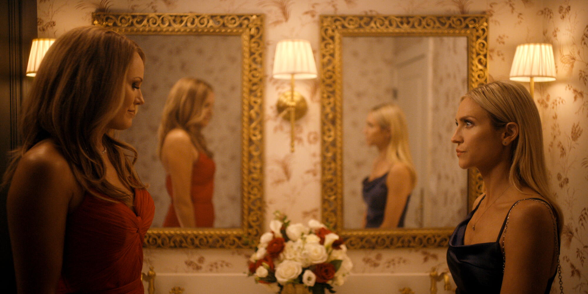 Two women in strappy dresses stand face to face in front of a pair of mirrors showing their reflection.