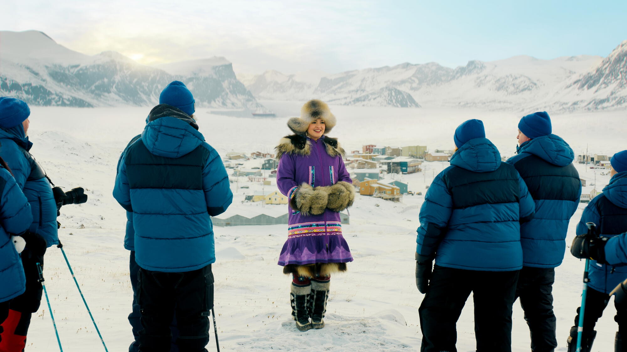A woman in a bright purple parka stands in front of a group of men in blue and black parkas on a snowy hilltop.