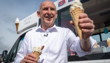 John Healey smiling, holding two ice cream cones, standing in front of an ice cream van.