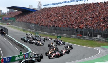 Formula 1 race cars on track with a packed grandstand of cheering fans at a major motorsport event.