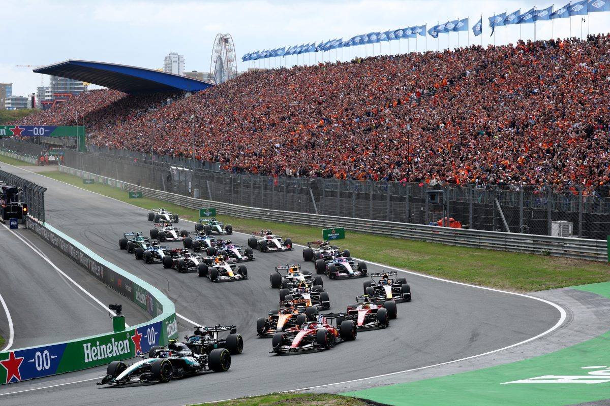Formula 1 race cars on track with a packed grandstand of cheering fans at a major motorsport event.