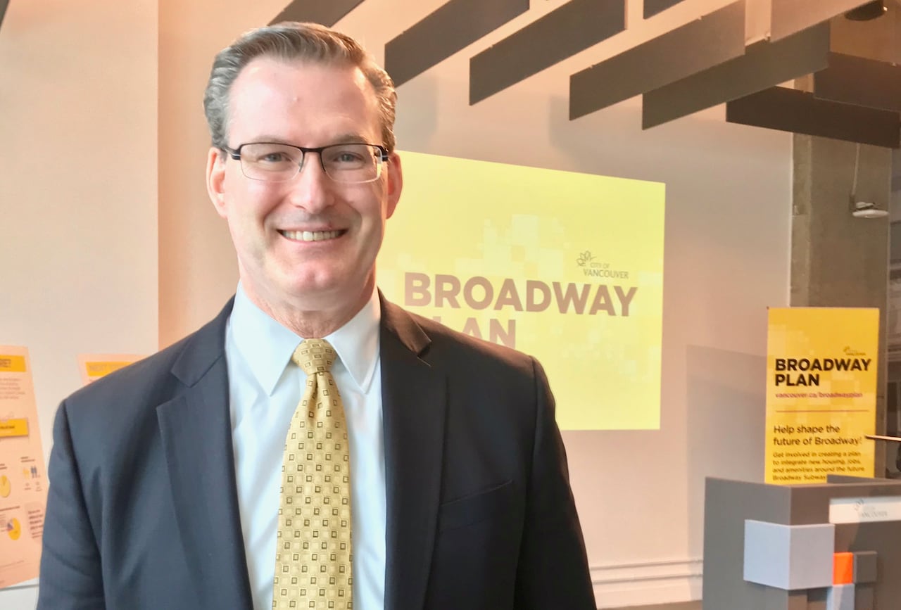 A white man smiles in front of a sign that reads 'Broadway Plan'.
