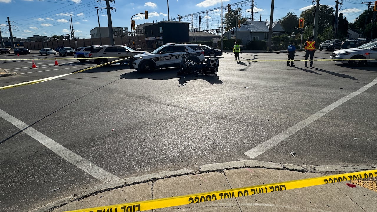 A city intersection is surrounded by yellow caution tape. A police vehicle and a motorcycle sit in the middle of the intersection