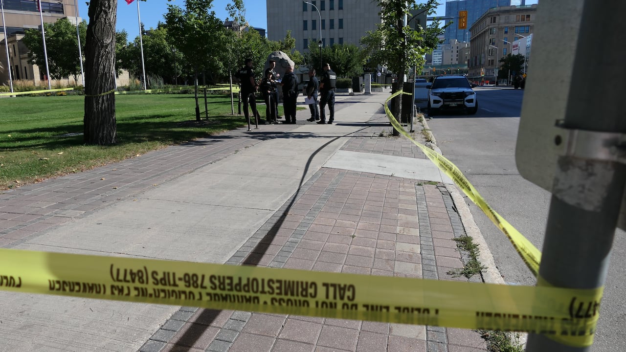 A Winnipeg police car is parked outside a stone building with spires and a red sign in front that reads "The University of Winnipeg." A section of sidewalk is blocked by police caution tape