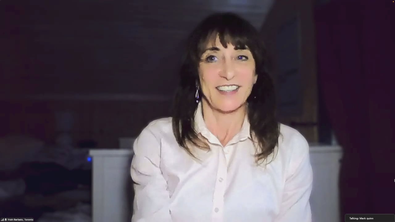 A woman with long, dark hair, wearing a white top speaks with a reporter with zoom