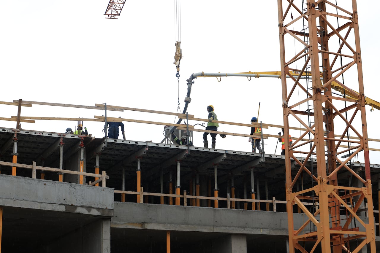 A construction worker in PPE leans on a temporary railing at the top of a building under construction. Other workers, also wearing PPE, are nearby.