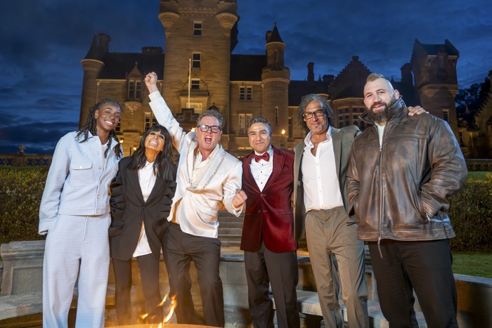 celebrity-traitors Cat Burns, Claudia Winkleman, Alan Carr, Nick Mohammed, David Olusoga & Joe Marler stood next to each other smiling ahead as Alan Carr raising one hand in the air.