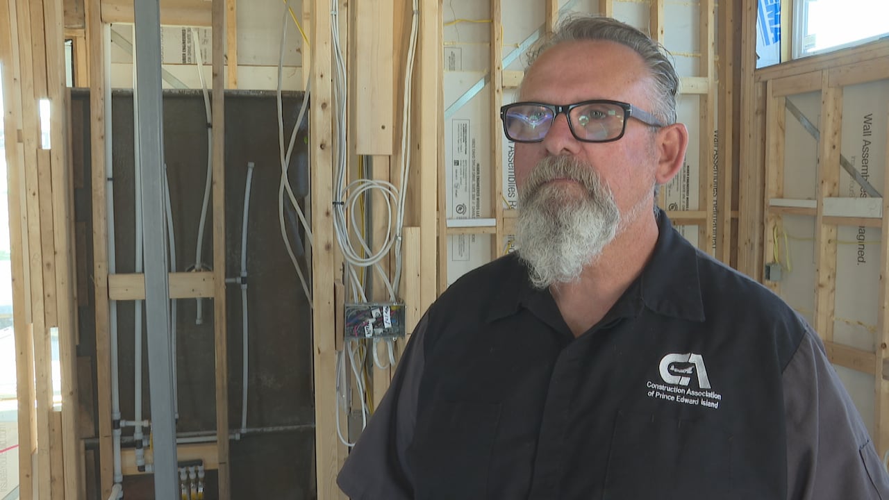 a man wearing glasses and a Construction Association of Prince Edward Island shirt inside an unfinished building framed with exposed wooden studs, wiring, and plumbing pipes.