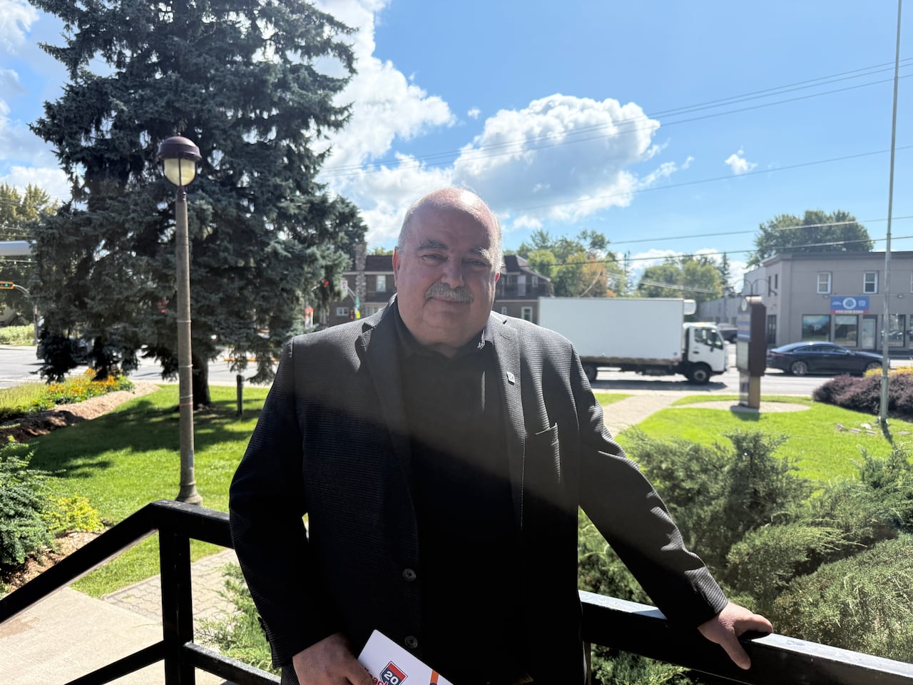 Pincourt mayor in black shirt and blazer stands outside with highway 20 in the background.