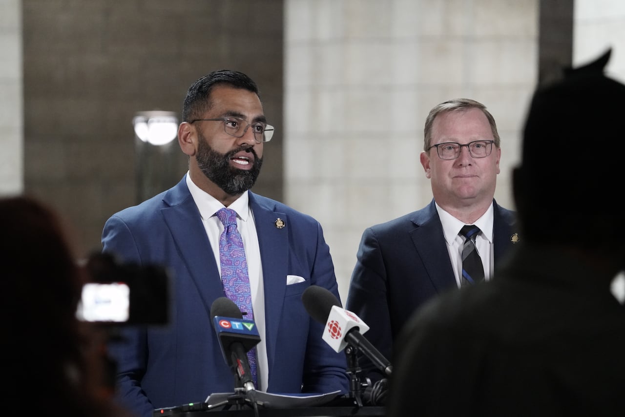 Two men in suits stand behind a lectern with microphones for an interview.