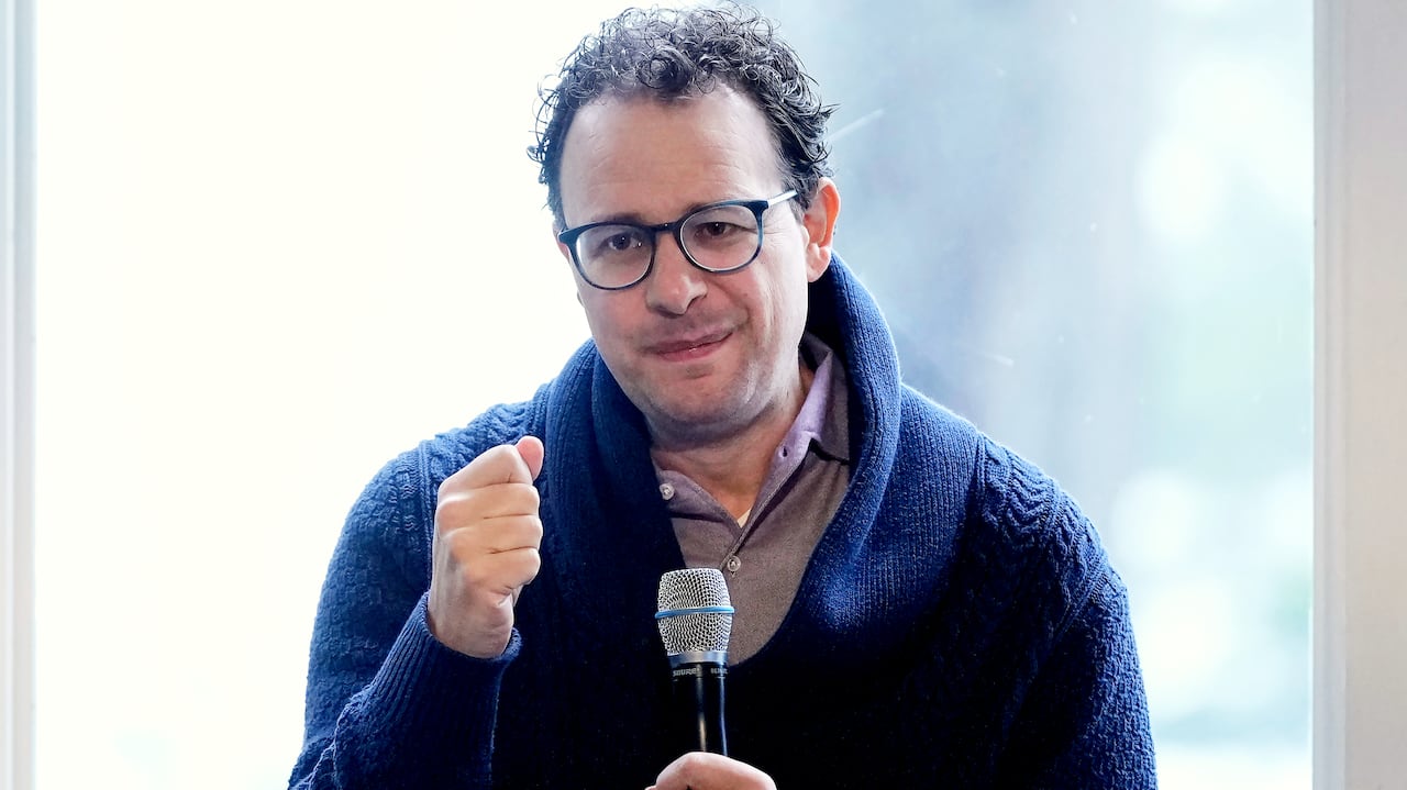 A man holds a microphone with his other fist held up, making a closed-mouth half-smile.