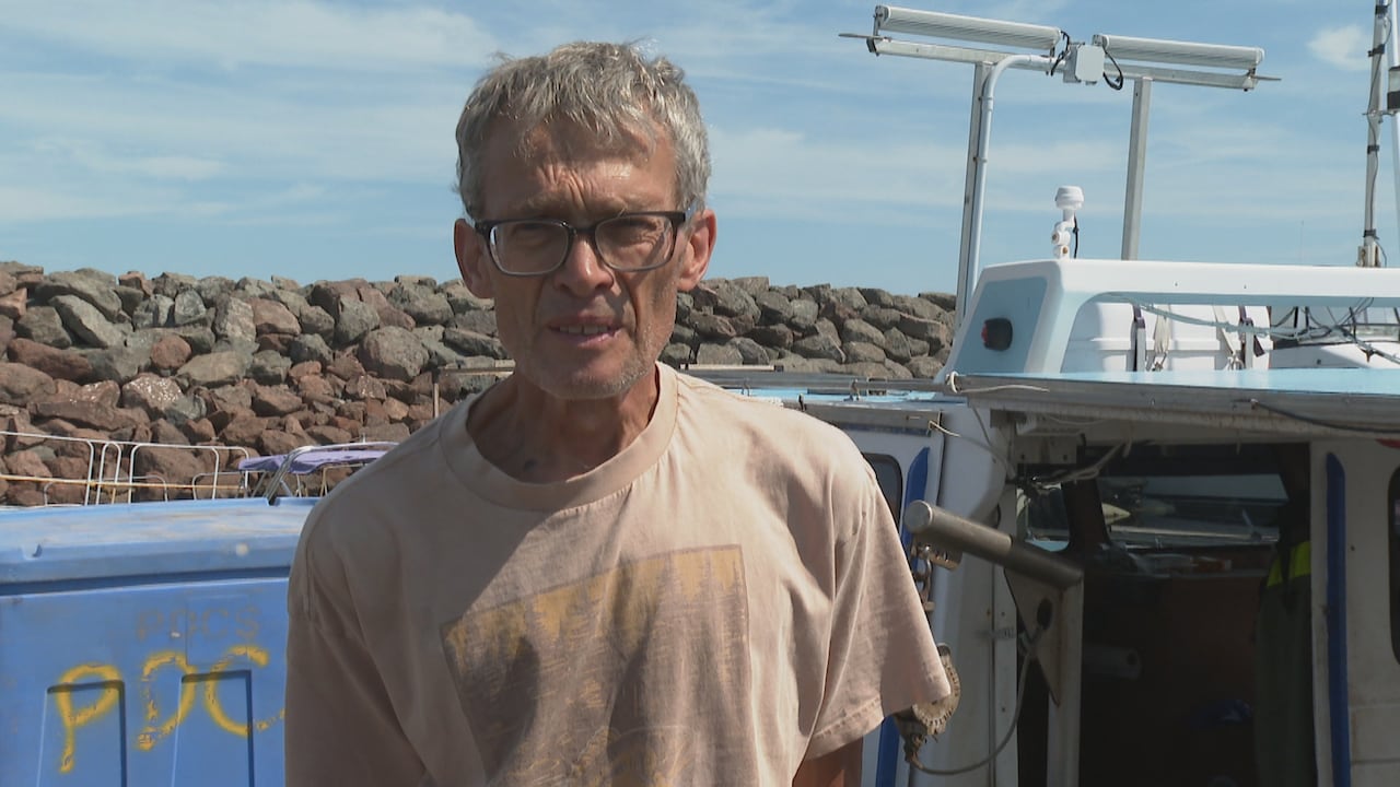 A man in his sixties with short gray hair, eye glasses and wearing a t-shirt, with parts of a boat cabin, large plastic bin and rock wall breakwater behind him.
