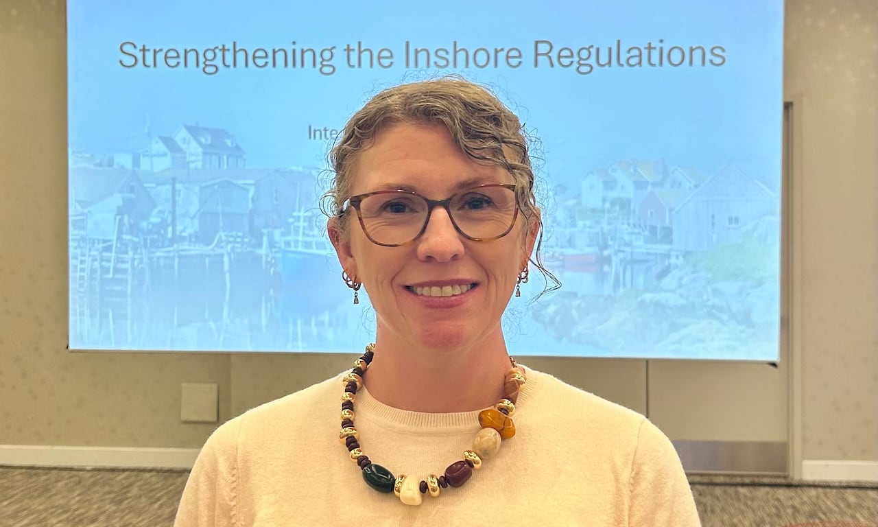 A woman wearing glasses, a beaded necklace and white t-shirt with her hair pulled back standing in front of a projection screen with a slide that says, "Strengthening the inshore regulations."