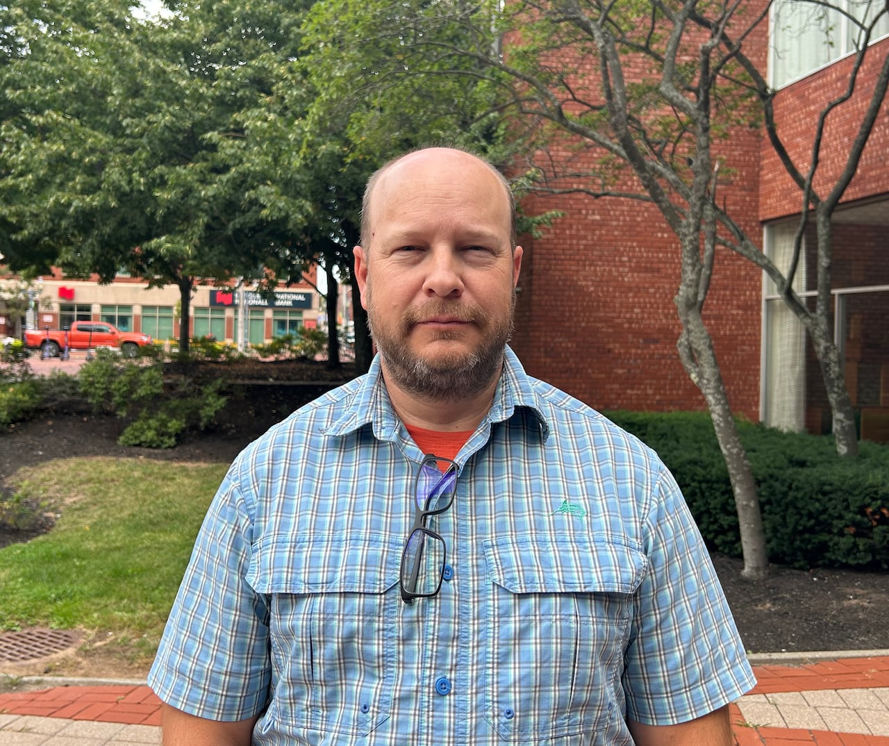 A man with a short beard wearing a short-sleeved blue checked shirt standing in a treed courtyard with a few vehicles visible in the background on a city street.