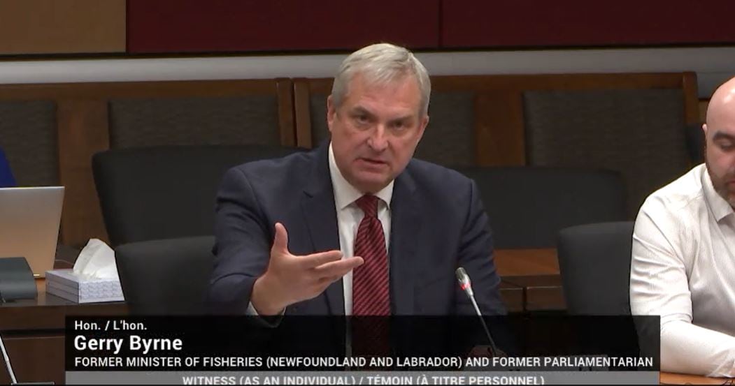 A man with gray hair and wearing a dark blue suit jacket, white shirt and maroon tie sits in the front row of a hearing room at a microphone, gesturing with one outstretched hand opened upward.
