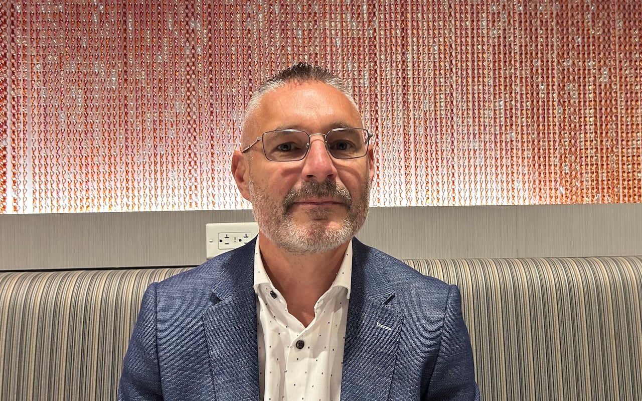 A man with short spiky gray hair and wearing glasses, a blue sports coat and white shirt with the top button open sits on a restaurant style bench with a cushioned back.