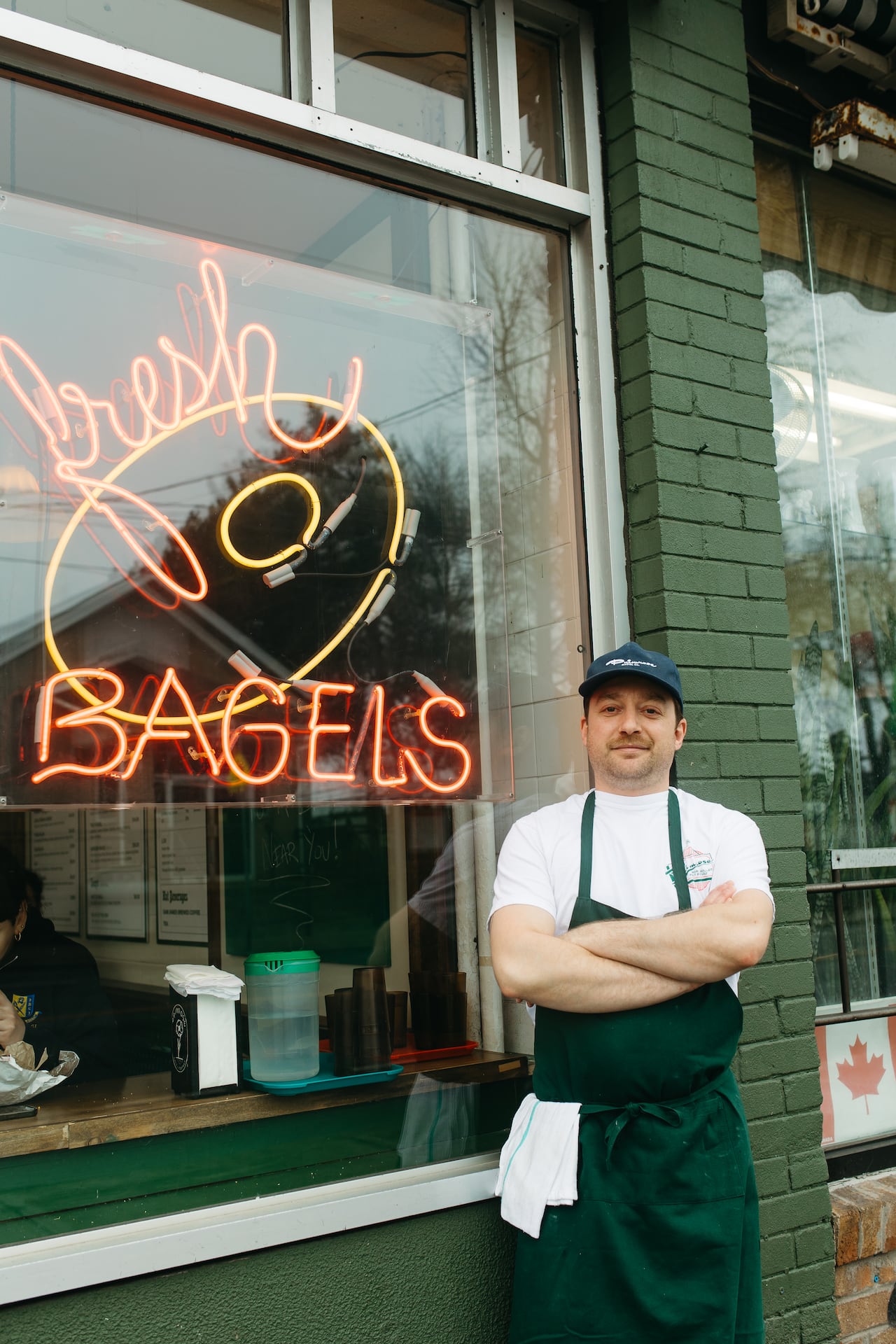 A man in an apron and baseball cap stands with his arms folded in front of a bagel store.