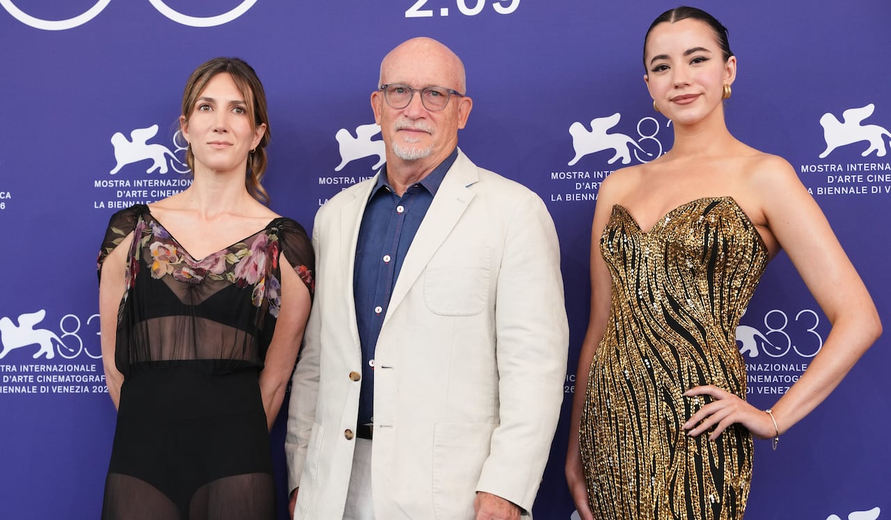 Three people stand together in front of a purple backdrop. Two women at the edges, with a man in a white suit in the middle.