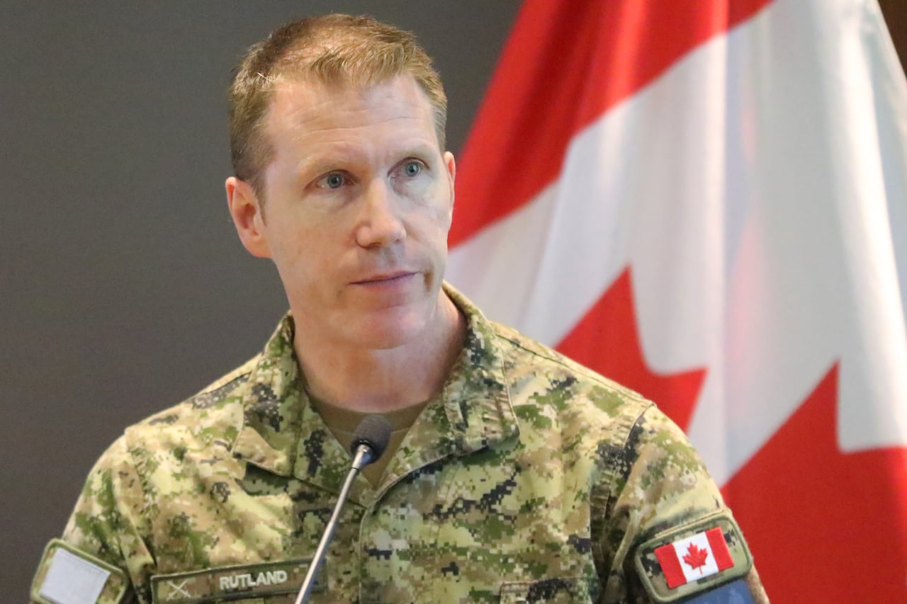 A man in a Canadian army uniform speaks into a microphone in front of a Canadian flag.