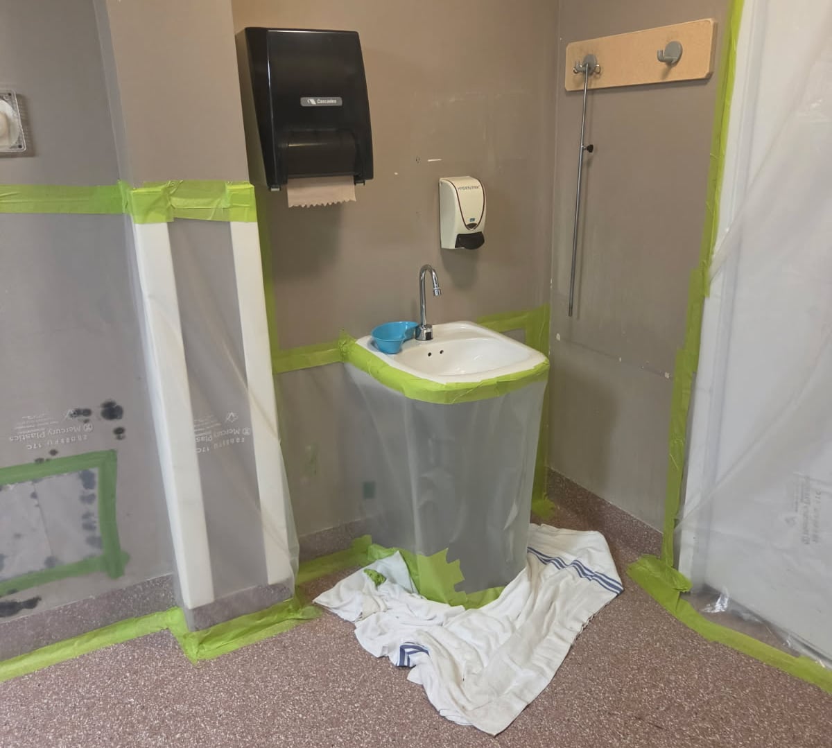 A sink, paper towel dispenser and door are taped off with plastic.