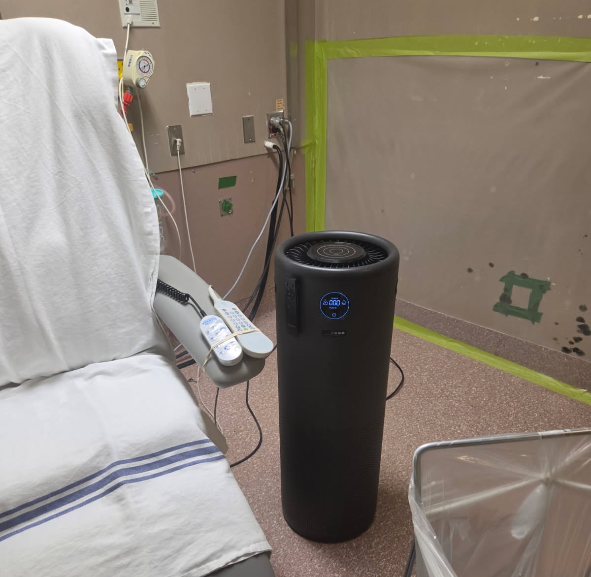 A black air purifier in the middle of a hospital room, beside a chair.