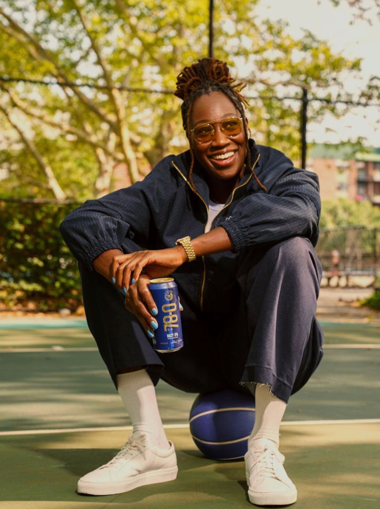 A smiling person wearing a black jacket and glasses, holding a blue beverage can, sitting on a basketball court surrounded by trees.