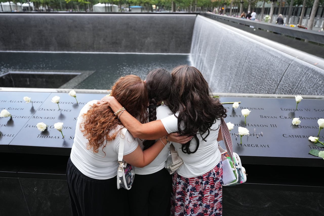 Three women are shown from behind, hugging in front of a concrete memorial with a water pool.