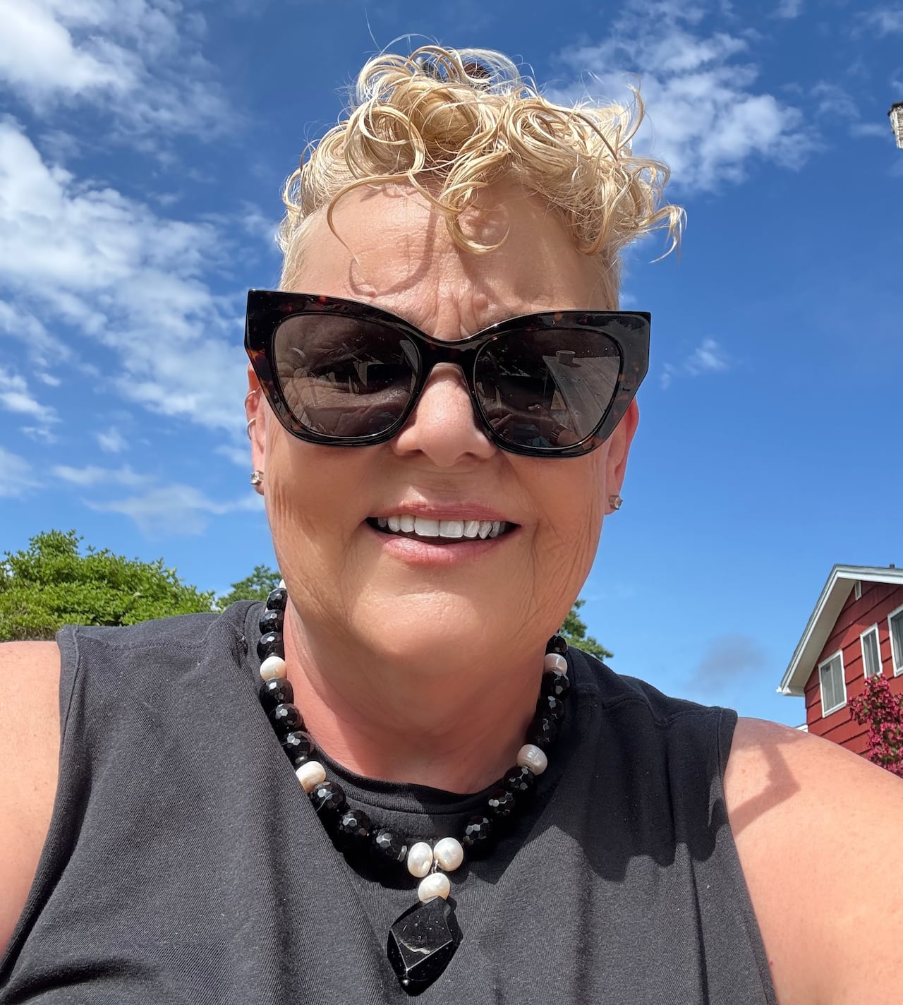A woman with blonde hair and large sunglasses takes a selfie, with a bright blue sky behind her.