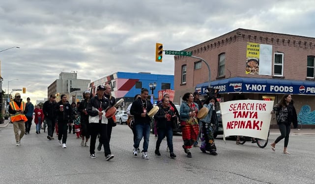 A group of people walk through the street past a city intersection holding a banner that reads "Search for Tanya Nepinak"