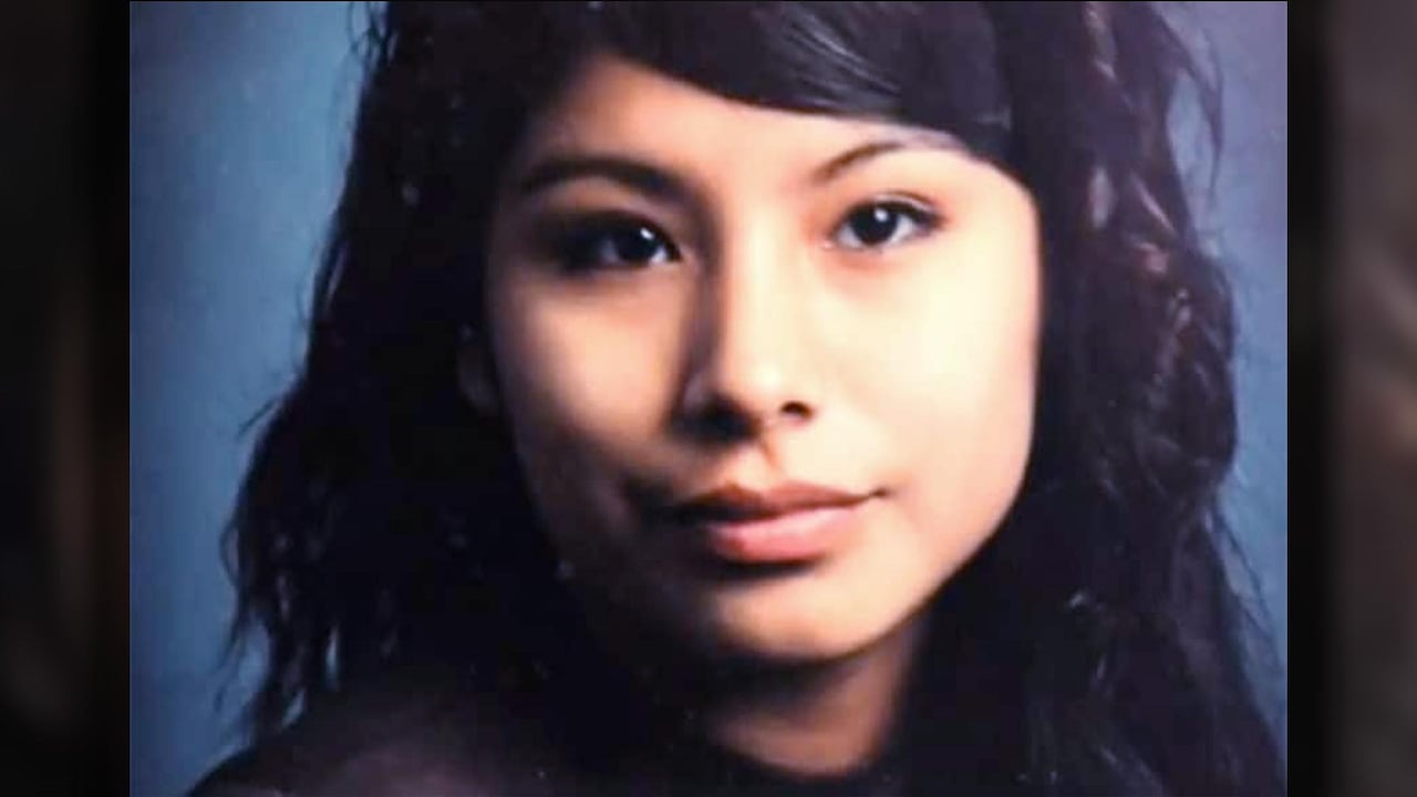 woman with dark hair and dark eyes looks straight to camera in front of a blue background