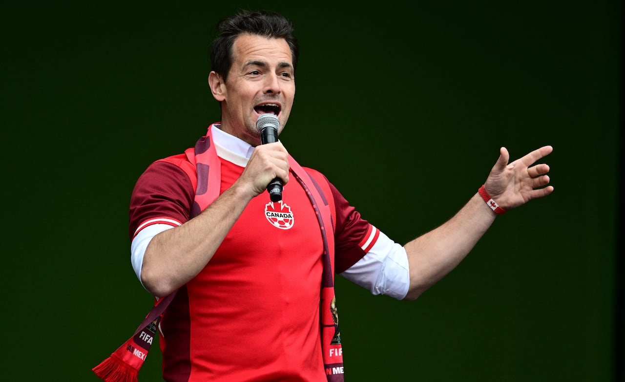 A man wearing a red shirt with a logo that has a soccer ball and the word "Canada" on it speaks into a microphone