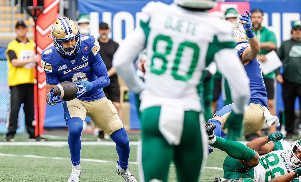 A football player in a blue and gold uniform that says "Blue Bombers" holds a football as a player in a white and green jersey runs toward him