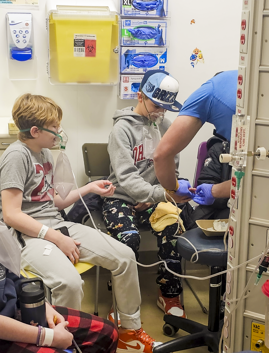 hockey players CO incident childrens hospital Two young hockey players wearing oxygen masks sit in a hospital treatment area as a healthcare worker administers care beside them, with oxygen tubing, medical supplies, and clinical equipment visible throughout the room following a carbon monoxide exposure incident.