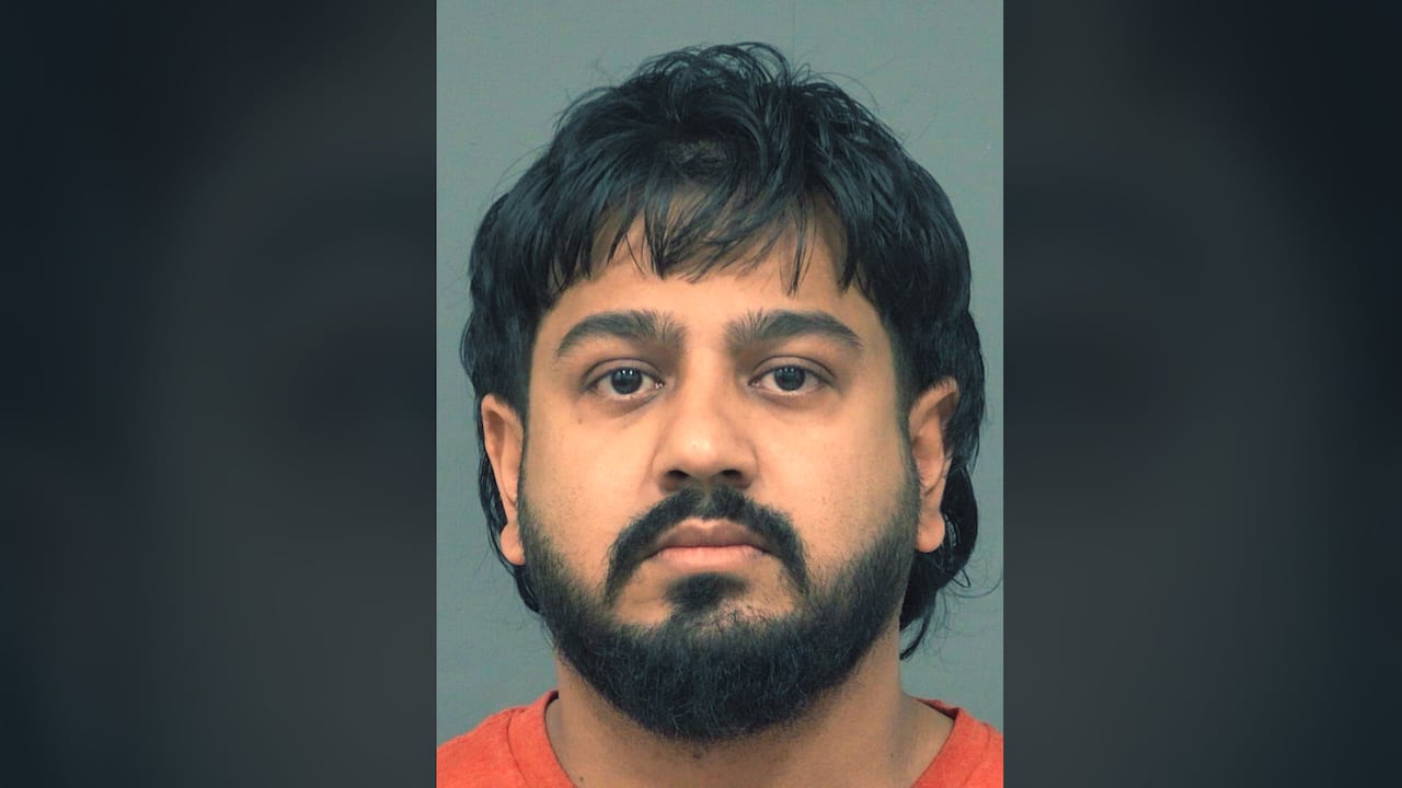 A mug shot shows a man with dark hair, a beard and mustache. He is wearing orange and looking into the camera. 