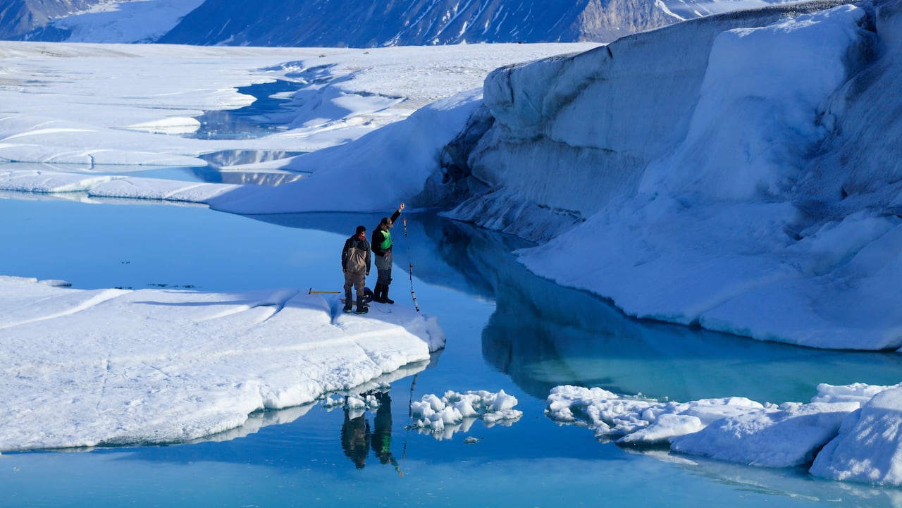Two men stand on a peninsula of ice and snow and hold a string into the water surrounding them
