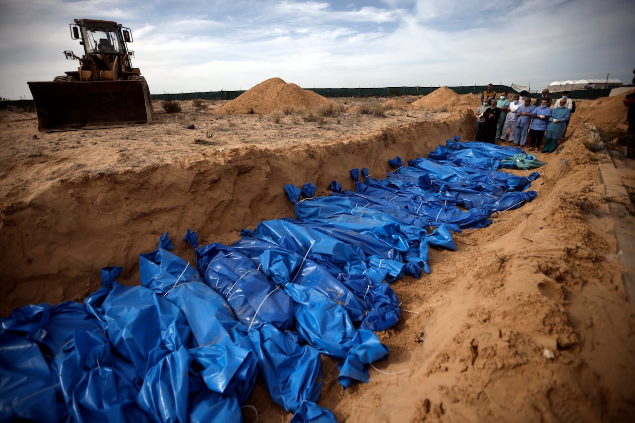 A line of bodies wrapped in blue plastic lie in a mass grave. A bulldozer and people who are praying are nearby.